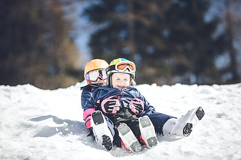 Zwei Kinder rodeln lachend im Schnee mit Helmen und Winterkleidung